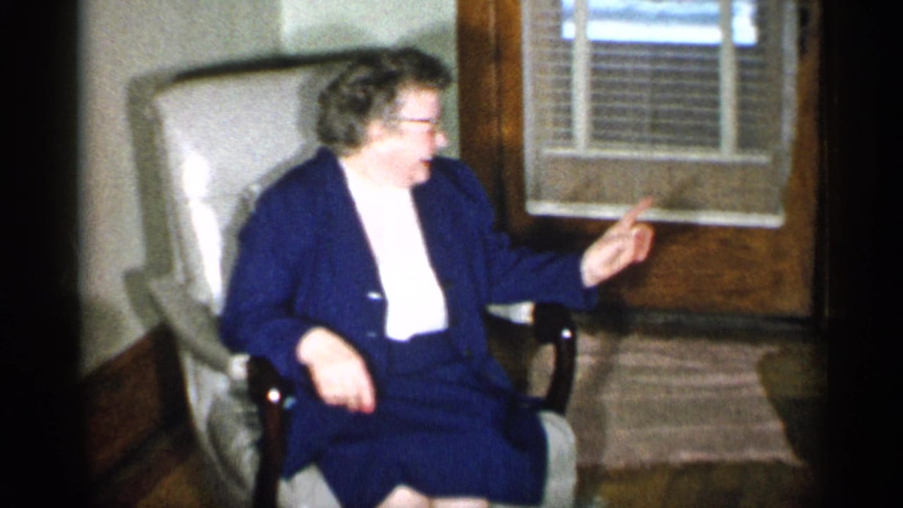 Older woman laughing and gesturing from a light armchair in a living room.