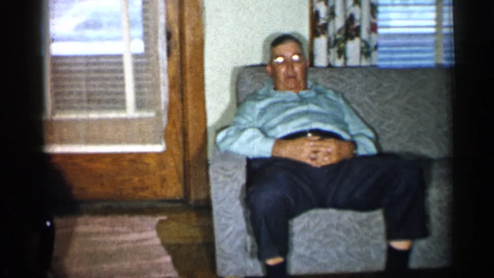 A man in a striped sweater appears to doze in a gray armchair inside the same private 1950s New York living room.