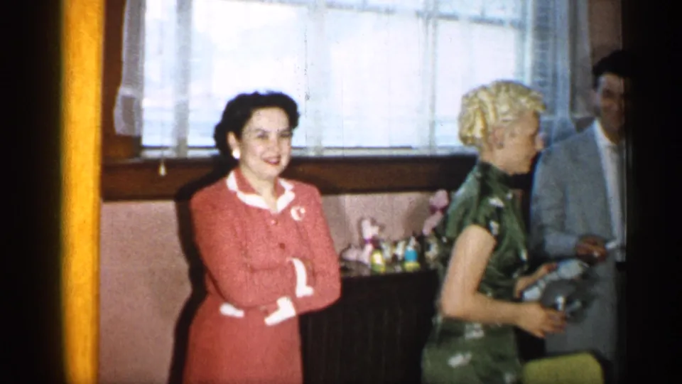 Several adults, including women in red and green outfits, smile and interact near a curtained window in a 1955 New York home interior.
