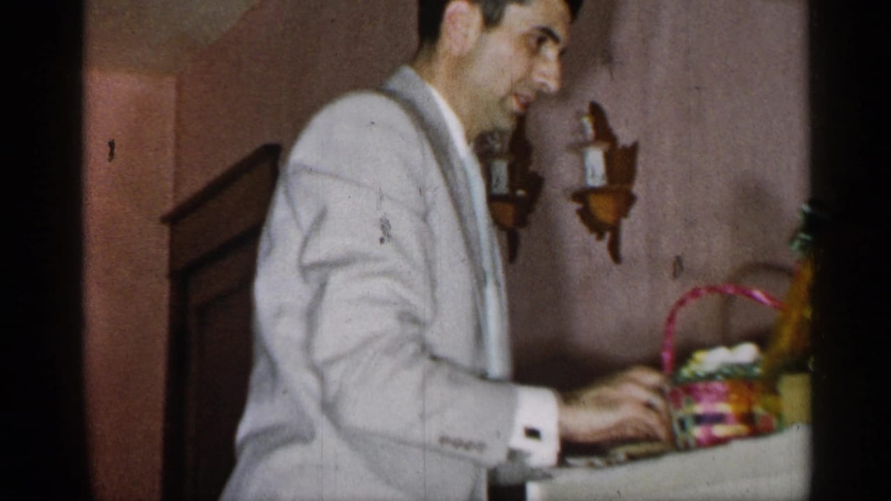In a 1955 New York home-movie frame, a man in a pale suit stands by a table and handles a colorful woven basket.