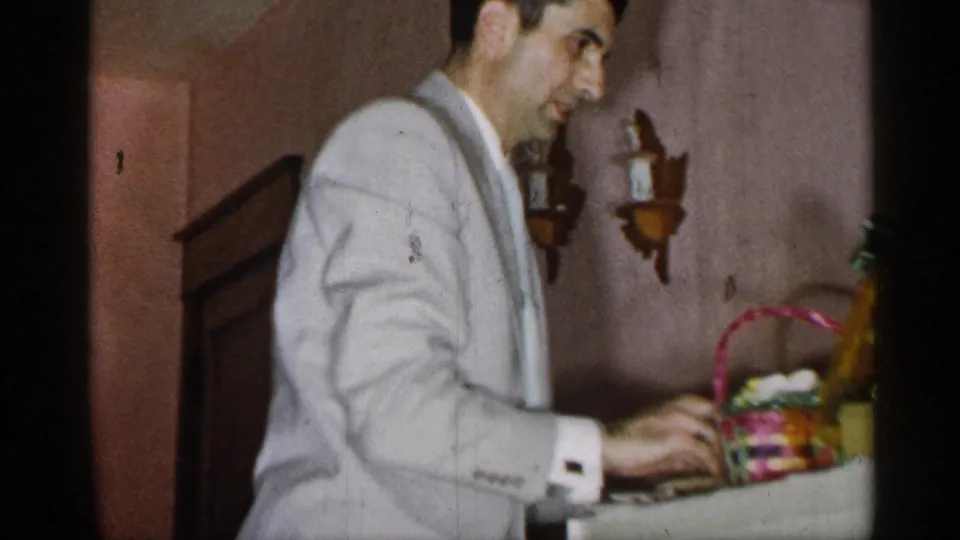 In a 1955 New York home-movie frame, a man in a pale suit stands by a table and handles a colorful woven basket.