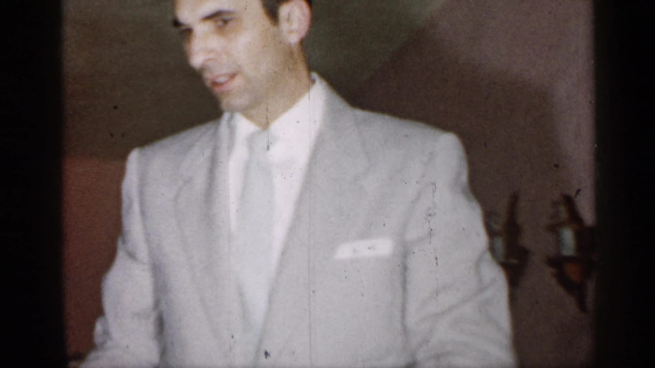 Man in a light suit faces near the camera in a warm interior.