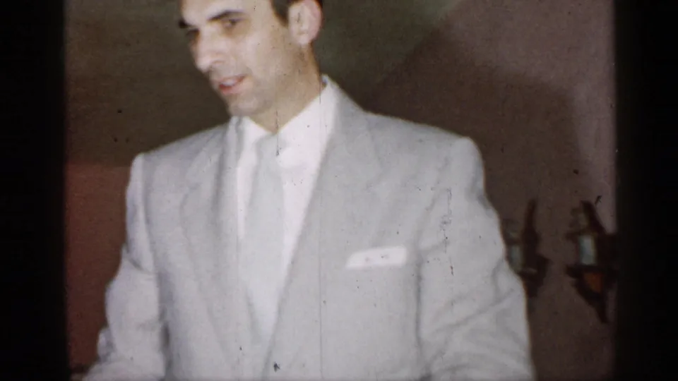 A man in a pale suit appears close to the camera inside a New York private interior, with wall sconces behind him.