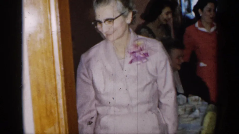 An elder woman in glasses, wearing a pink suit and floral corsage, smiles in a crowded 1955 New York indoor gathering.