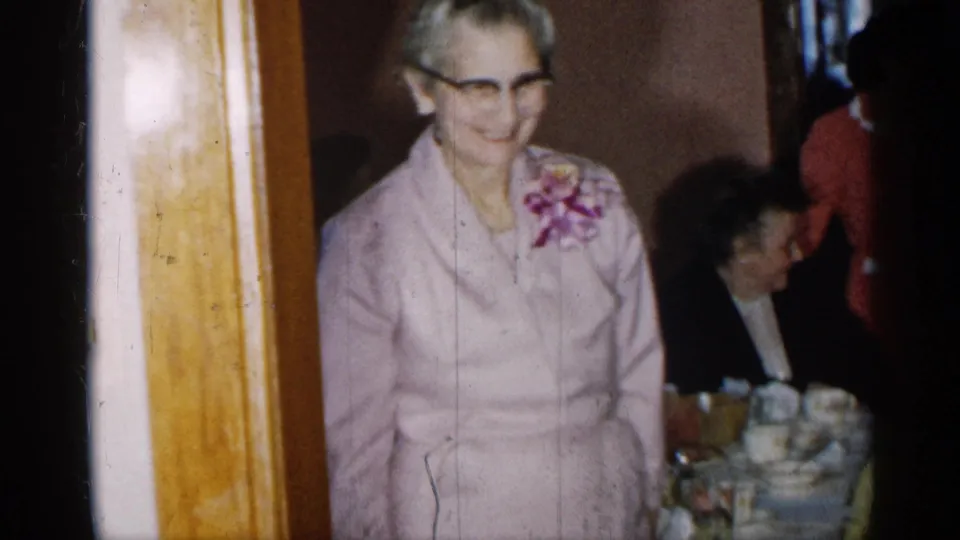 The elder woman in a pink suit smiles beside a table set with cups, with other guests visible in the private New York interior.