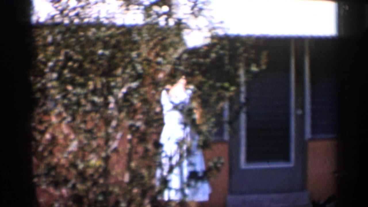 1958 Alabama frame of a woman in a light dress near a garden wall and exterior doorway, with foliage filling much of the background.
