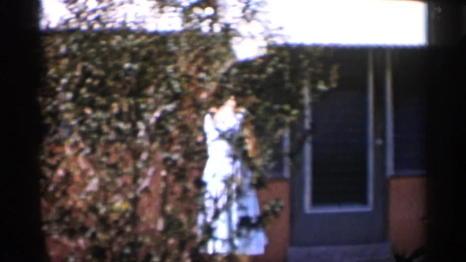 1958 Alabama frame of a woman in a light dress near a garden wall and exterior doorway, with foliage filling much of the background.