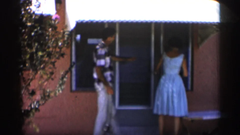 1958 Alabama home-movie frame showing a man in patterned shirt and a woman in a light dress near a storefront or building entrance.