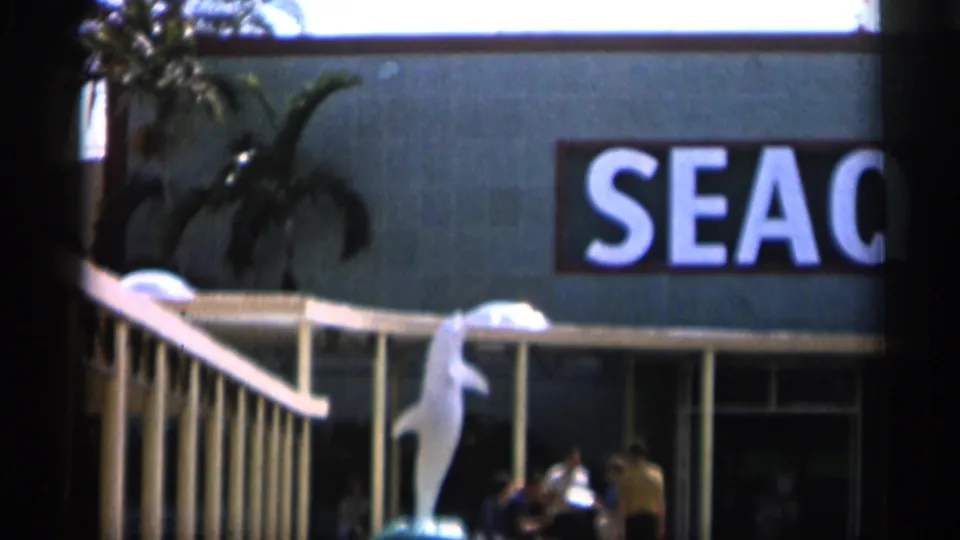 1958 Alabama frame showing a partial SEAQ storefront sign, exterior railing, plants, and figures near the building entrance.