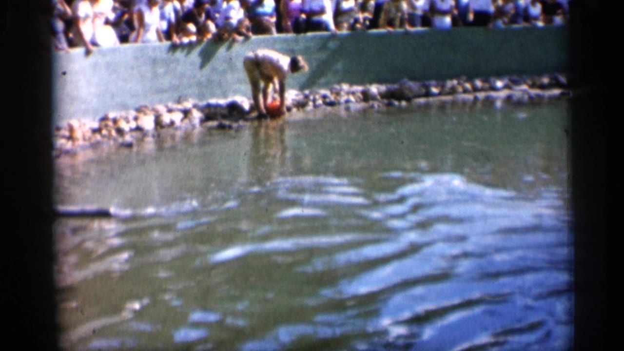 1958 Alabama home-movie frame of a keeper bending at the pond edge as visitors watch from behind a green zoo barrier.