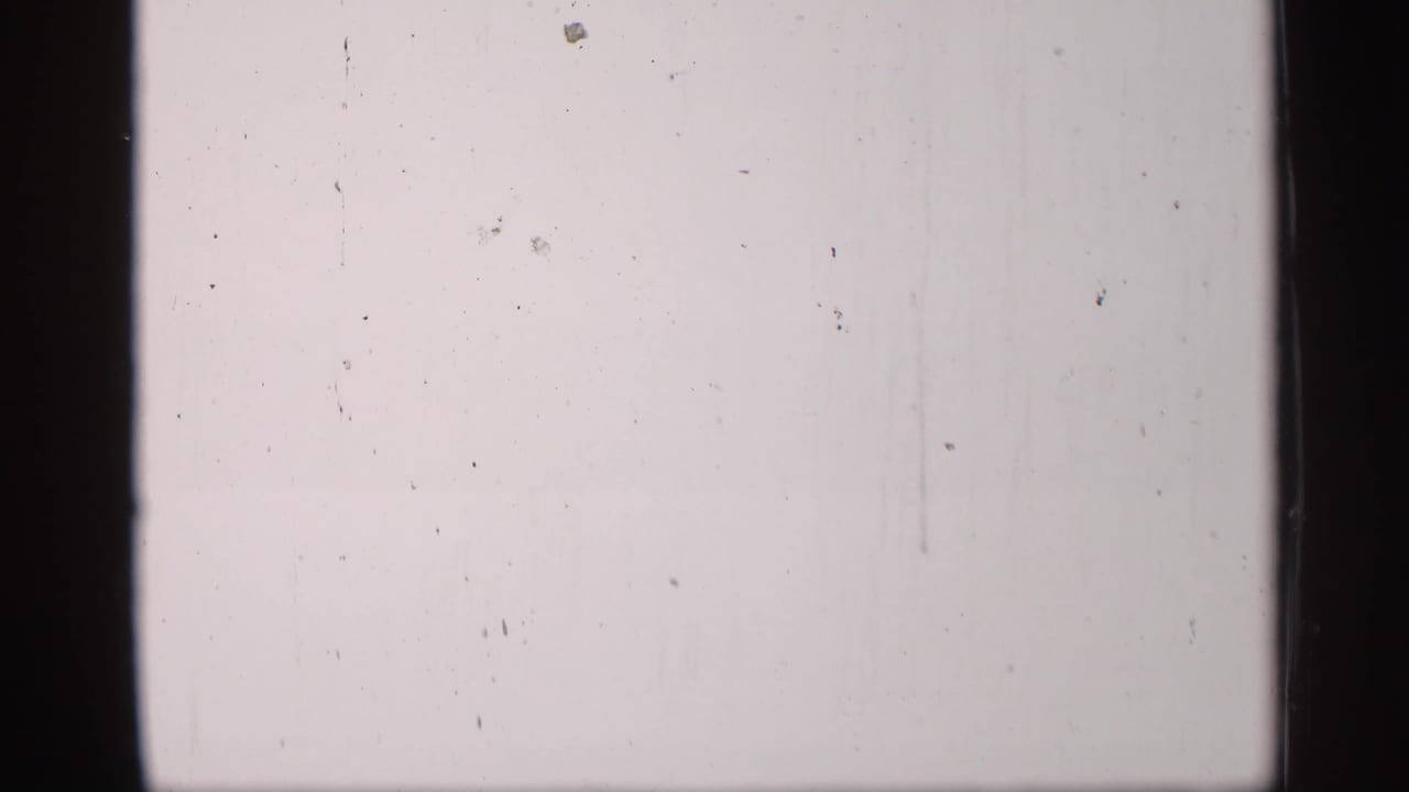 Pale overexposed 8mm frame with visible dust, scratches, and dark vertical borders, with little scene detail visible.