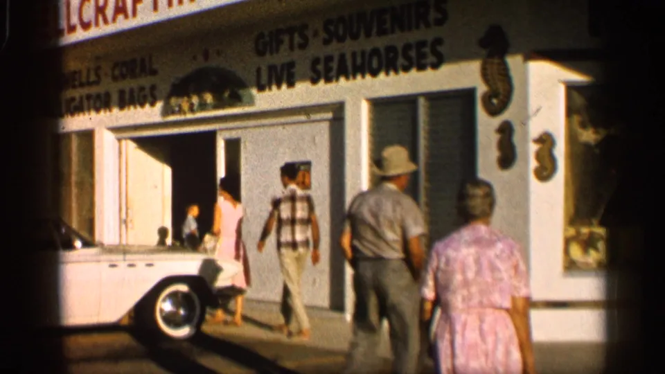 1958 Alabama home-movie frame showing adults outside a souvenir shop with signs for gifts and live seahorses.