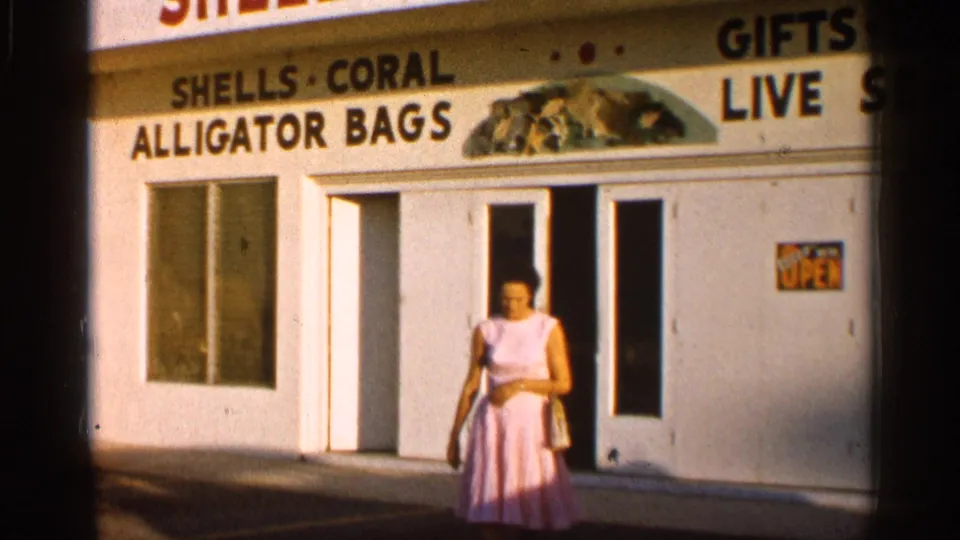 1958 Alabama 8mm frame showing a woman in a pink dress passing a souvenir shop with shells, coral, alligator bags, and gifts signage.