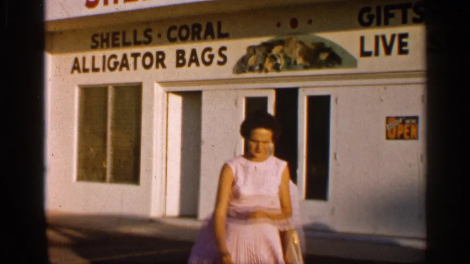 1958 Alabama 8mm frame with a woman walking close to camera outside a souvenir shop advertising alligator bags, gifts, and live sea items.