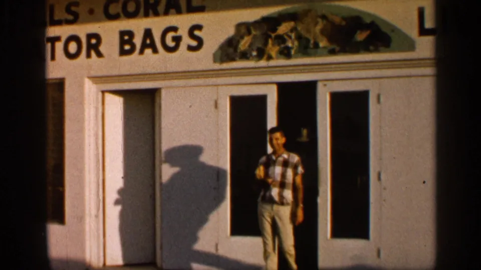 1958 Alabama home-movie frame of a man leaving a tourist souvenir shop with visible signage for coral, alligator bags, and other goods.