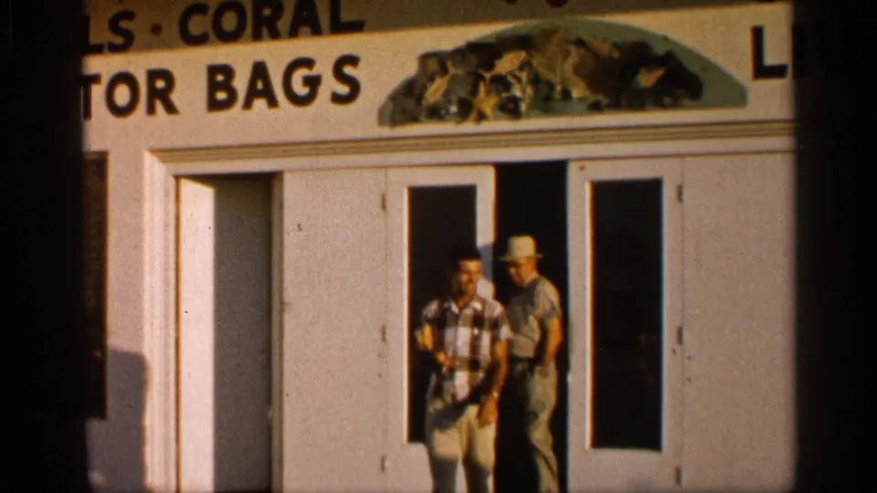 In 1958 Alabama footage, two men exit a storefront with glass-panel doors and a sign mentioning shells, coral, and bags.