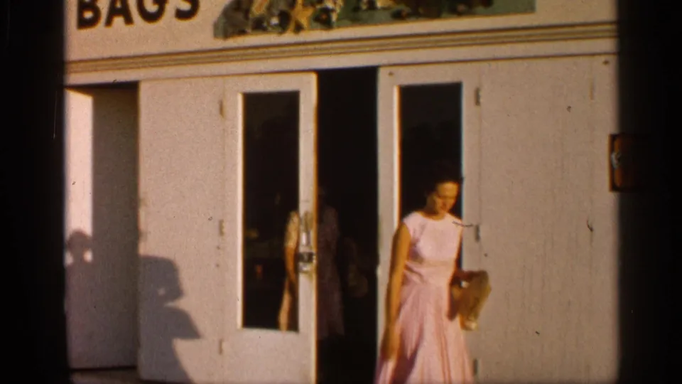 A woman in a light pink dress steps out of the same Alabama storefront, carrying a handbag or small parcel in bright sun.