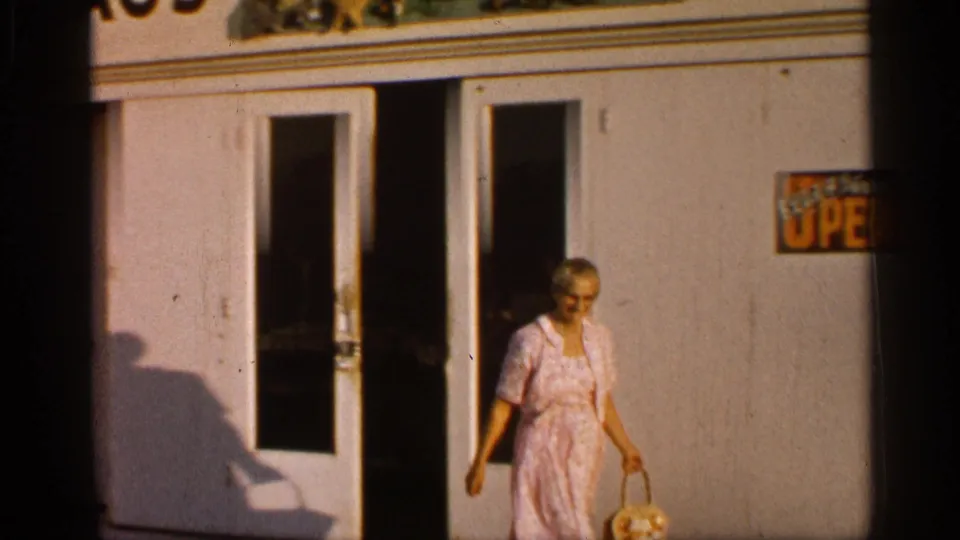 In Alabama 1958 home-movie footage, a woman in a pink dress exits a white building with a basket near a partly visible sign.