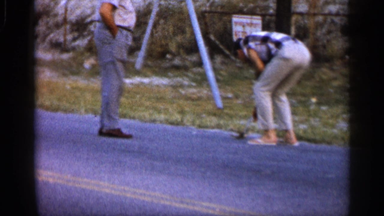 Two men are beside an Alabama roadside in 1958 footage, with one bending near a pole or post while the other stands watching.