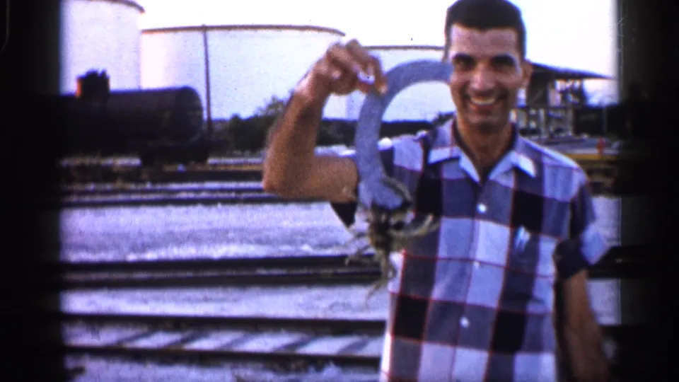 A smiling man in a plaid shirt holds up a crab near rail tracks, tank cars, and water in 1958 Alabama home-movie footage.