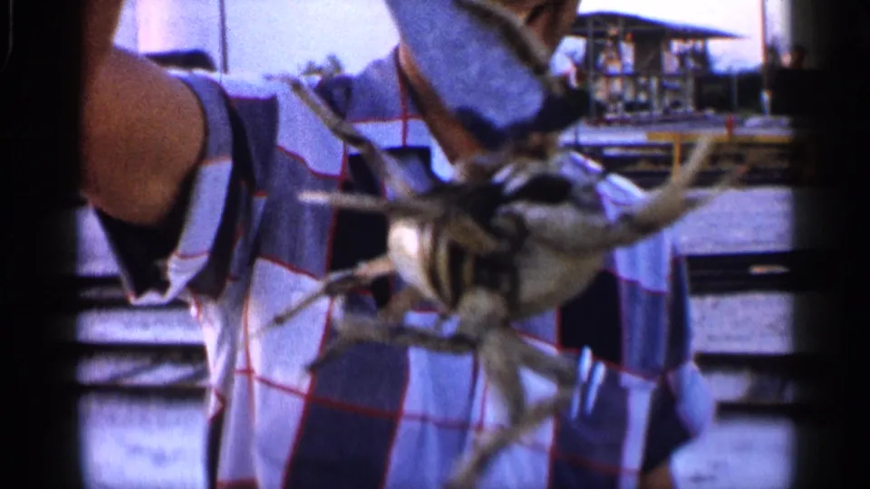 Close-up of a crab held toward the camera, partly obscuring a man in a plaid shirt near rail-side water in Alabama footage.