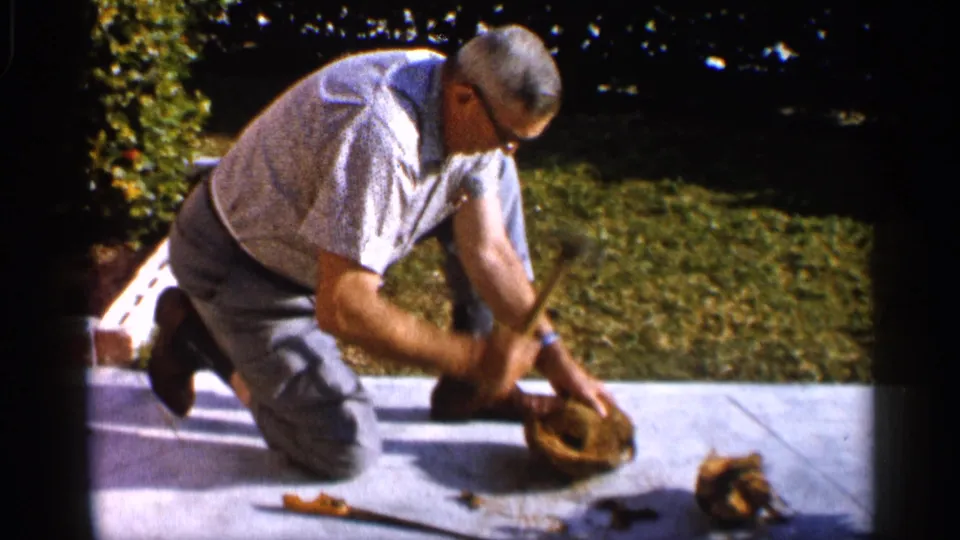 In a 1958 Alabama yard scene, a man in glasses kneels on a concrete sidewalk near a hammer and small broken material.