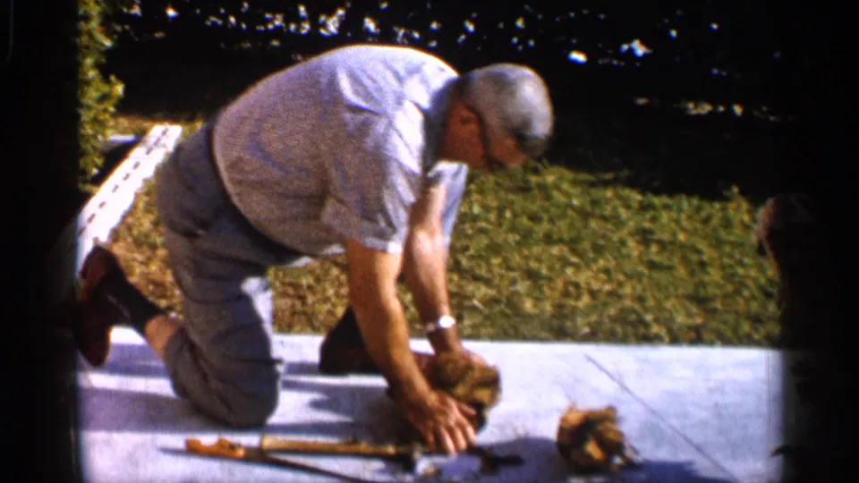 The man bends over the concrete sidewalk in the Alabama home-movie scene, reaching toward material near a hammer.