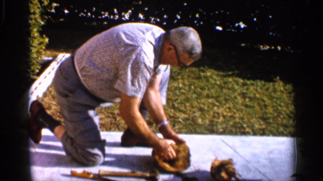 Man kneels on a concrete patio while working on a coconut with tools nearby.