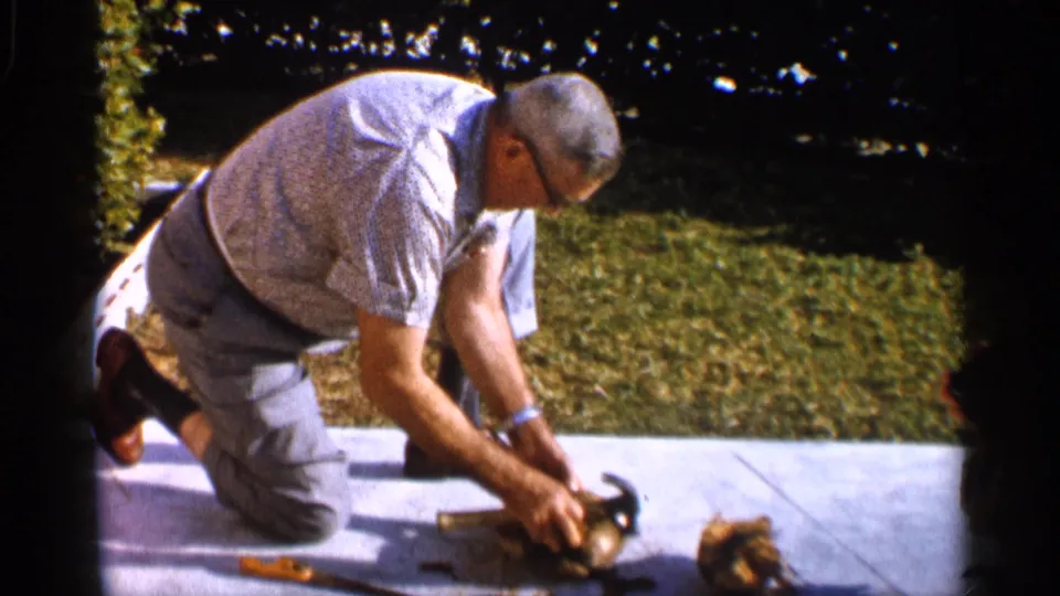 A man in glasses steadies a coconut on an outdoor patio, with hand tools and shell pieces visible nearby.