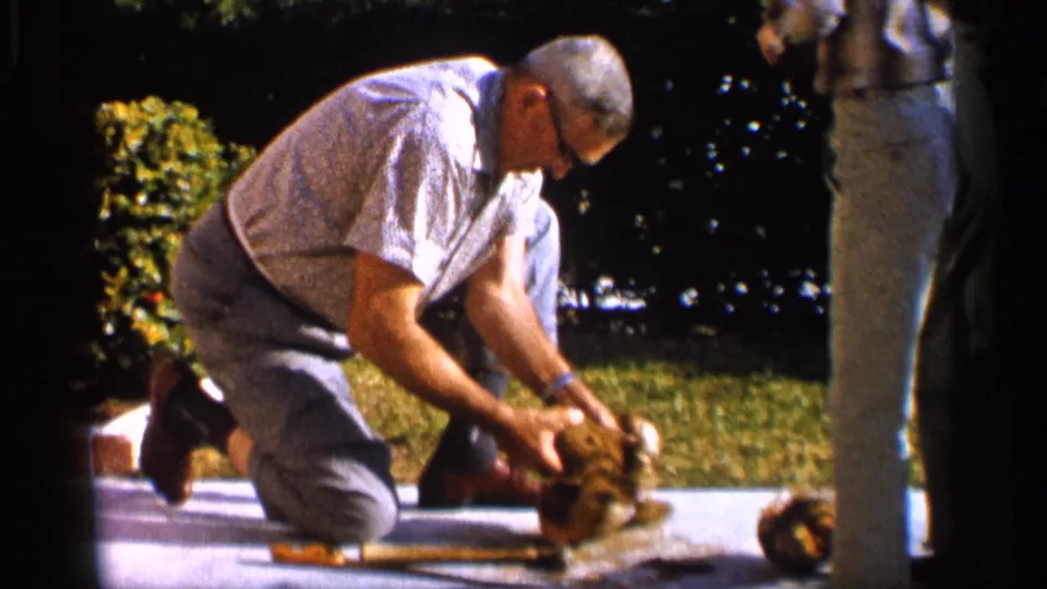 The kneeling man continues opening the coconut as another adult stands partly visible at the right edge of the 1958 Alabama frame.