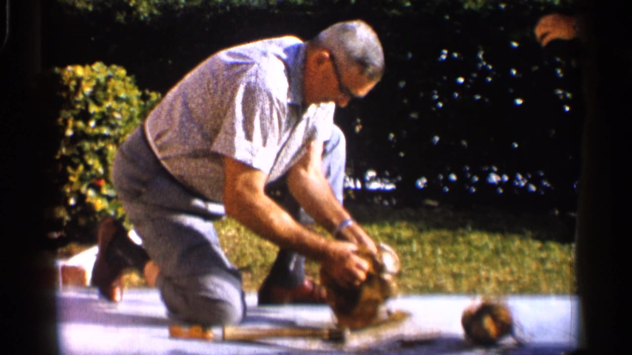 Man with glasses kneels on a driveway while holding a coconut near a tool.