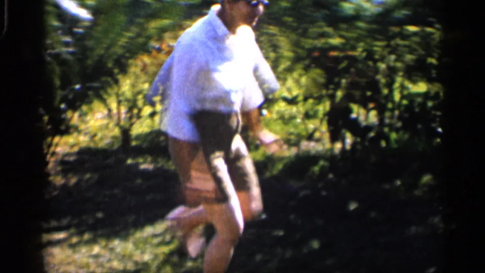 The man stands in the sunny yard holding the coconut, while a second person is partly visible near the edge of the 8mm frame.
