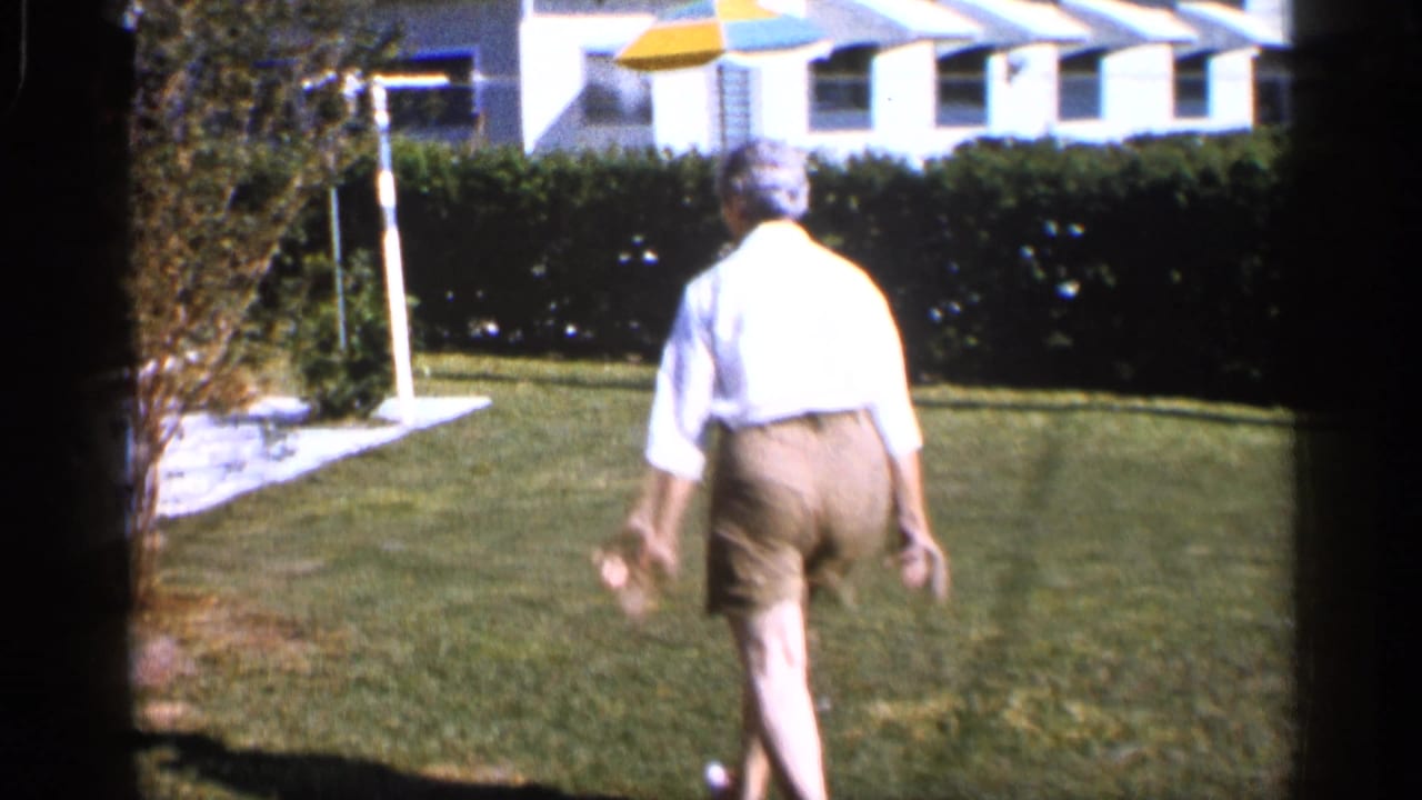 Barefoot man walks away across a lawn toward a hedge and house.