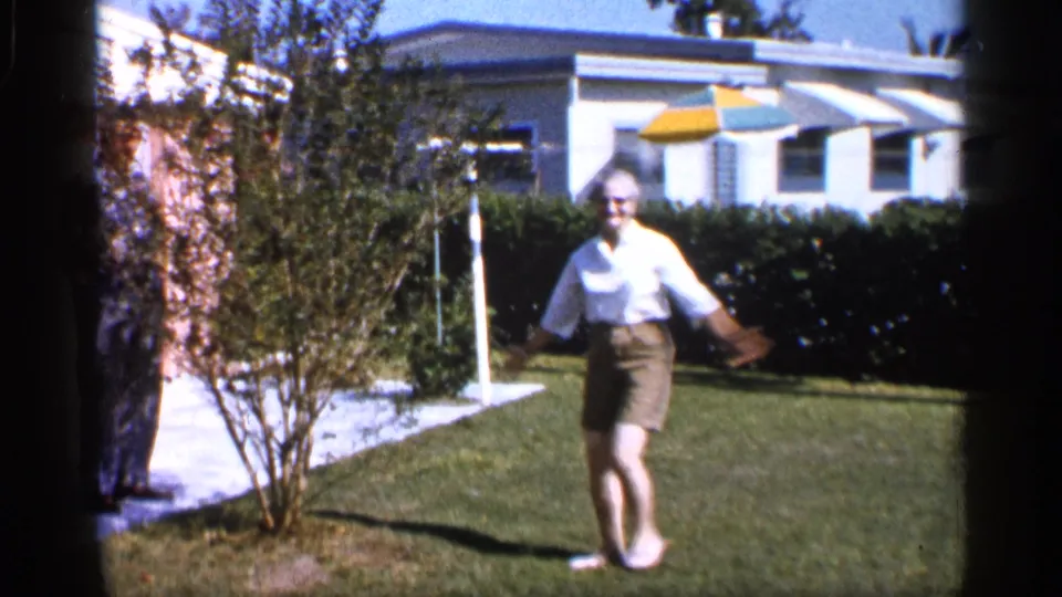The barefoot man turns toward the camera on the Alabama lawn, arms out, with a hedge, shrub, house, and striped umbrella behind him.