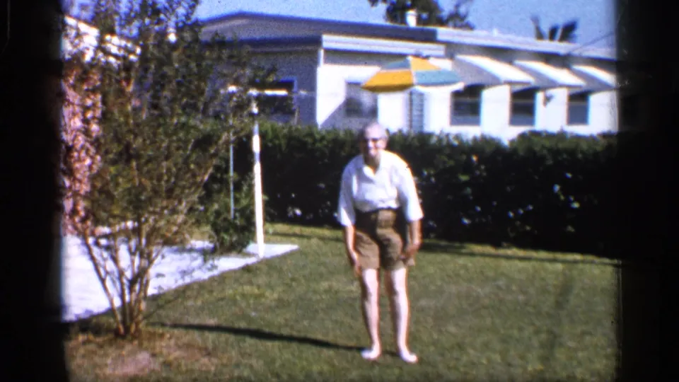The man stands facing the camera in the 1958 Alabama yard, with trimmed grass, hedge, house windows, and striped umbrella visible behind him.