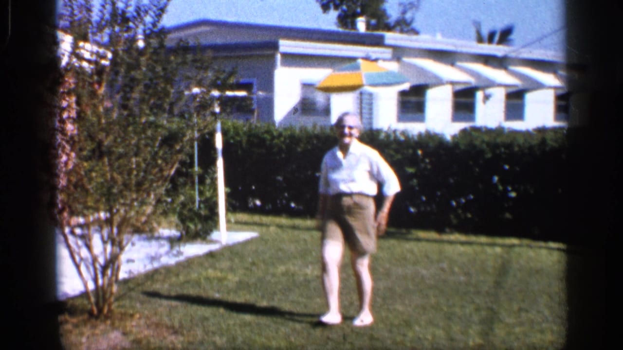 Woman in a white shirt and khaki shorts stands barefoot on a lawn near a hedge and white house.