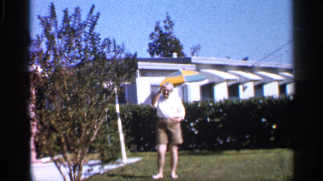 The woman waves toward the camera in a bright Alabama yard, with a hedge, white house, awnings, and patio umbrella behind her.