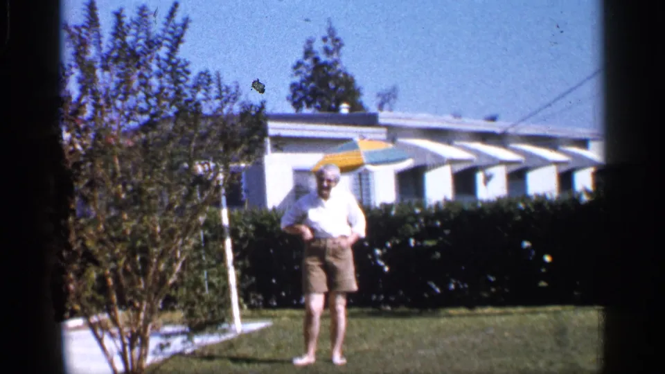 A smiling woman stands barefoot in an Alabama yard, framed by a small tree, trimmed hedge, striped awnings, and a white house.