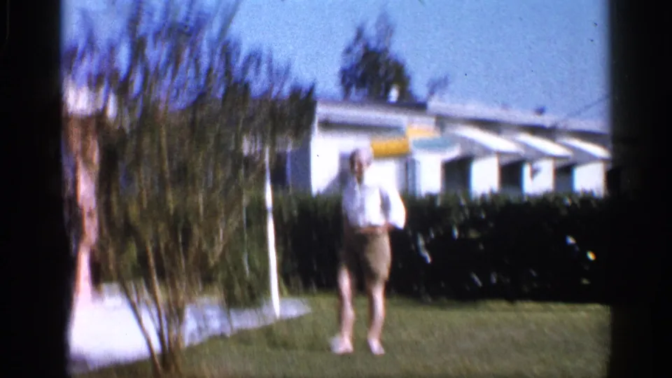 A soft, slightly blurred 8mm frame shows the woman in the yard before the hedge, white house, striped awnings, and umbrella.