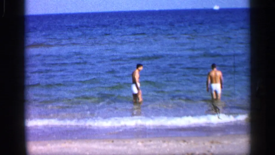1958 Alabama-linked beach frame showing two men in light swim trunks standing in shallow surf under a blue sky.