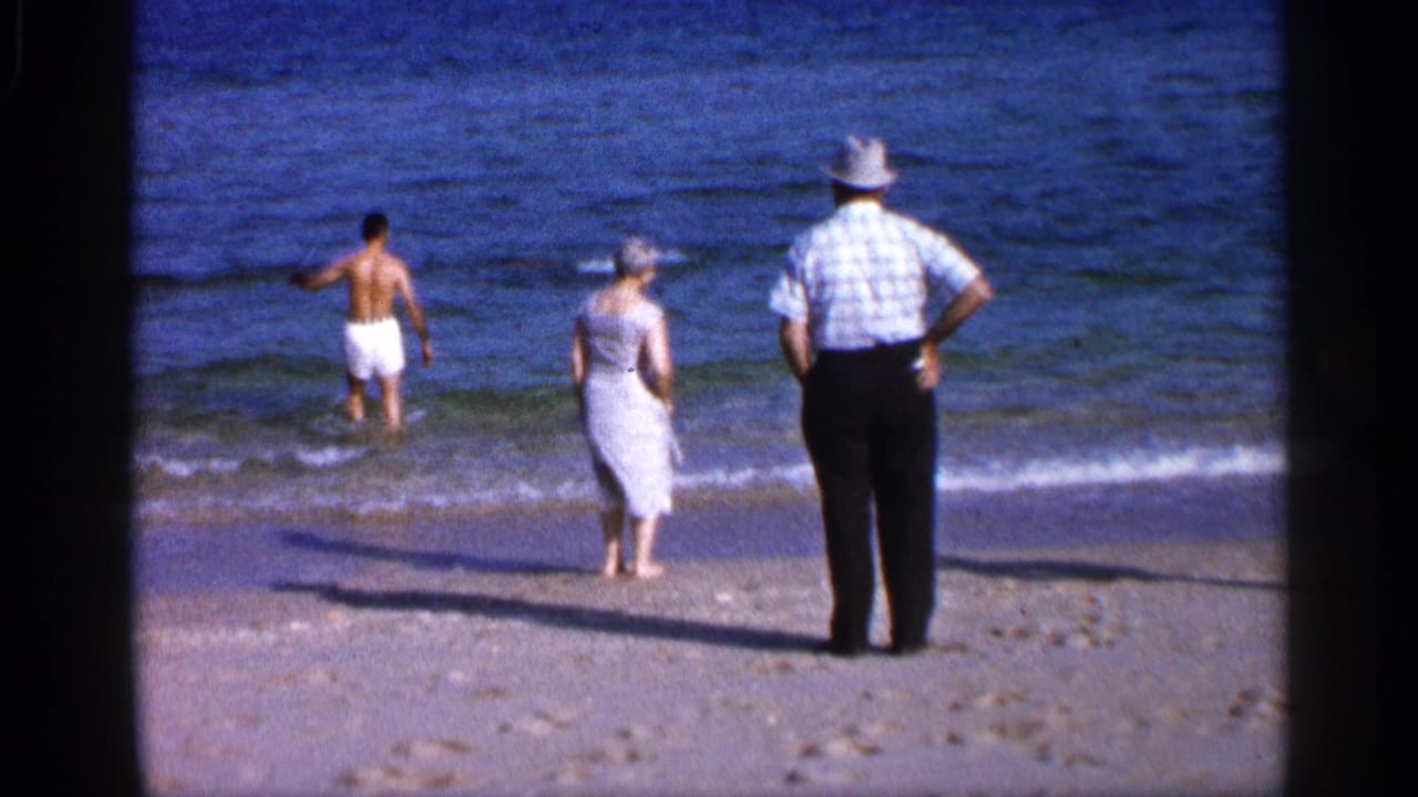 Man in white swim trunks wades into surf while two adults stand on the sandy shore.