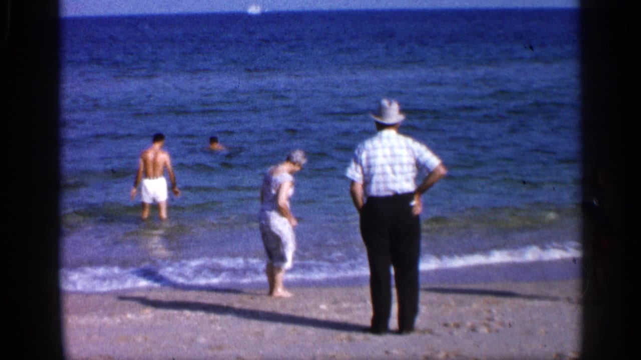 1958 Alabama beach frame with a man in white trunks near the waves, a woman stepping along shore, and an older man in a hat watching the water.