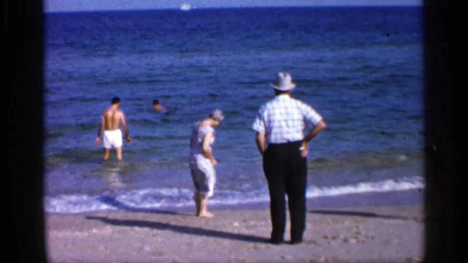 1958 Alabama beach frame with a man in white trunks near the waves, a woman stepping along shore, and an older man in a hat watching the water.
