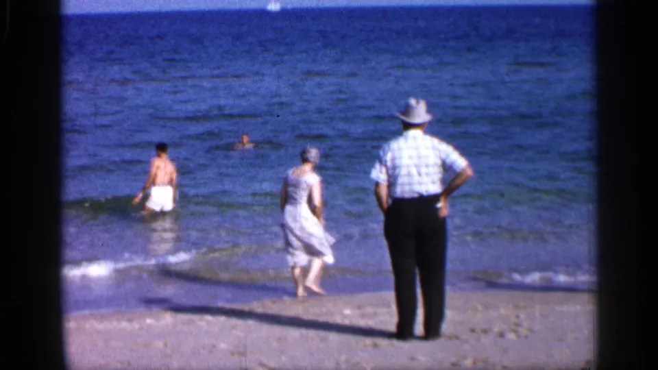 Color 8mm frame from the Alabama coast showing a woman in a light dress and a man in a hat watching as another man stands in shallow surf.