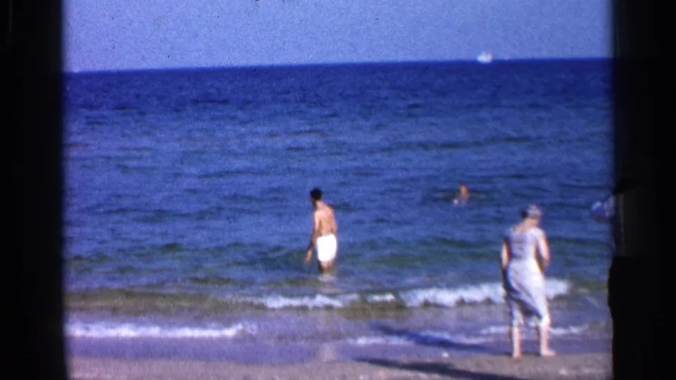1958 Alabama coastal frame showing a man in white trunks in the surf, a woman in a light dress at the shoreline, and distant swimmers under a bright sky.