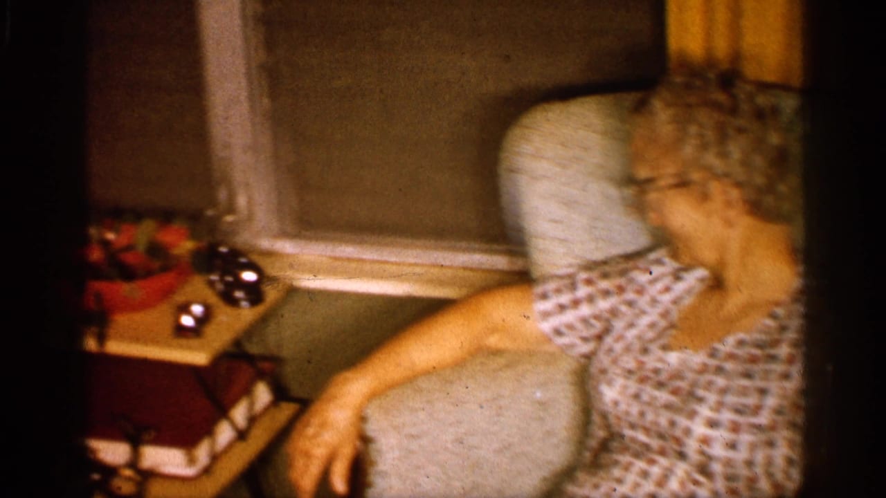 Older woman in glasses and a patterned dress sits in an armchair inside a 1958 Alabama family living room.