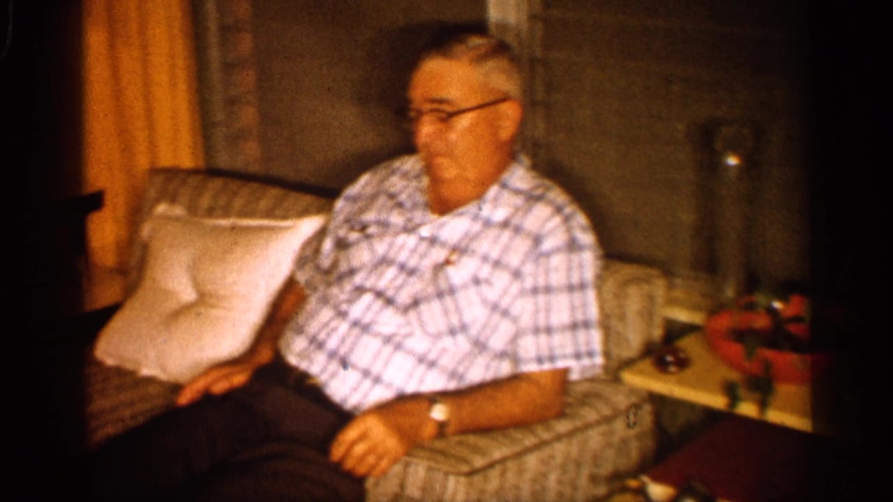 Older man seated on a sofa with pillows and a side table nearby.
