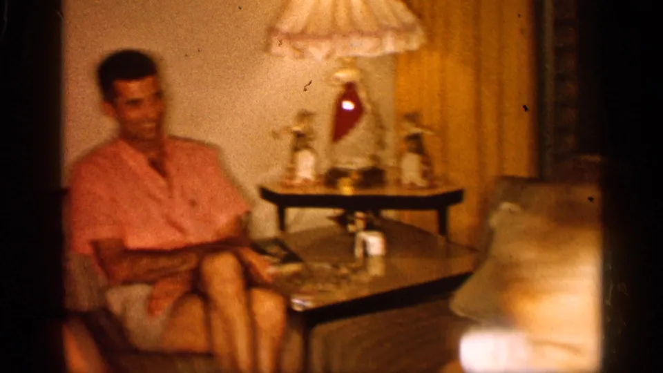 Adult man sits in a living-room chair near a coffee table and fringed lamp in 1958 Alabama home-movie footage.