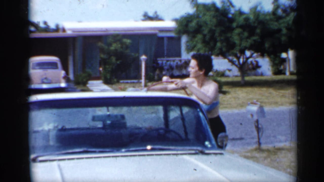 Woman washing a light-colored car in a suburban driveway with houses and a vintage sedan behind her.
