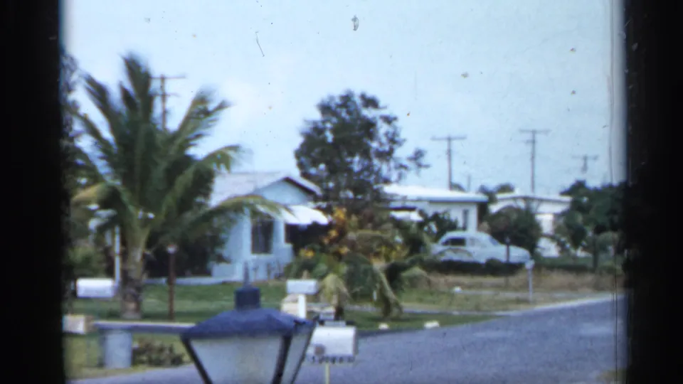 Wide residential street view in 1958 Florida footage, with palms, mailboxes, low houses, lawns, and a parked car near a driveway.